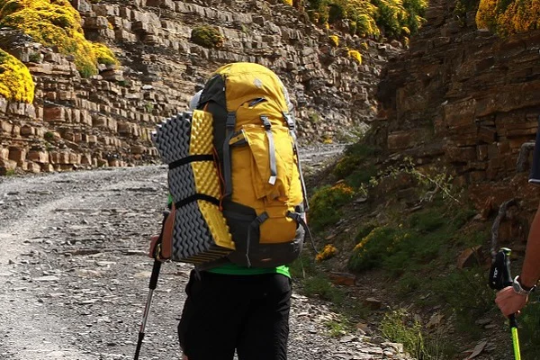 Person mit gelbem Rucksack wandert einen Kiesweg hinaut, am Rucksack ist eine gelbe Isomatte außen befestigt.