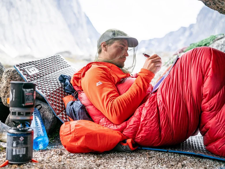 Outdoor Imagebild - Person liegt im freien auf einer Isomatte in einem roten Schlafsack. Sie trägt eine Kappe mit Moskitonetz, außerdem zu sehen eine Trinkflasche und ein Outdoorkocher mit Kartusche.