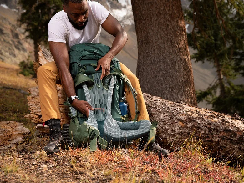 Person sitzt auf einem Baumstamm und hat einen grünen Rucksack. Sie verstellt den Rückenträger des Rucksacks.