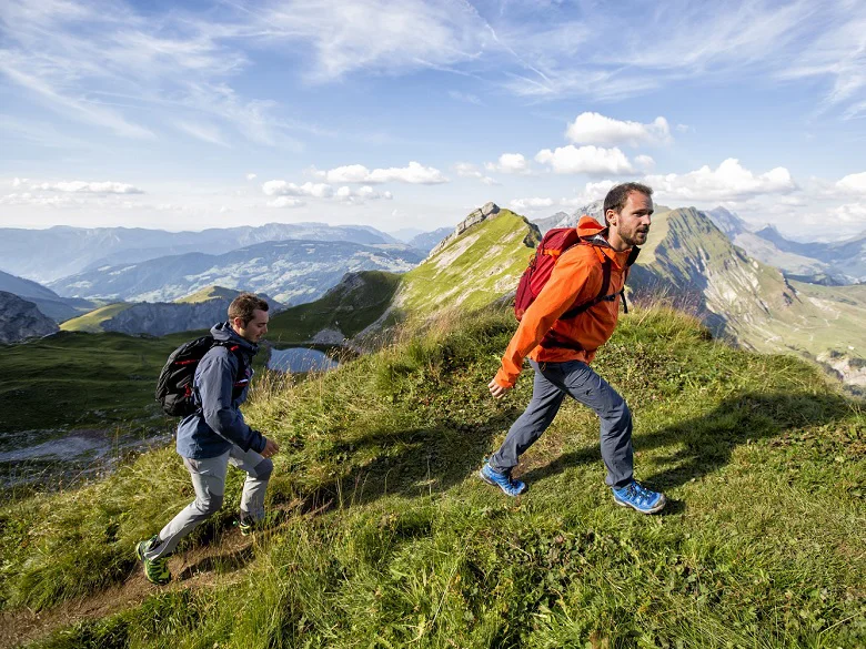 Outdoor Imagebild - zwei Personen wandern im Gebirge über das Gras. Sie sind mit Funktionsbekeldung und Wanderrucksack ausgestattet.