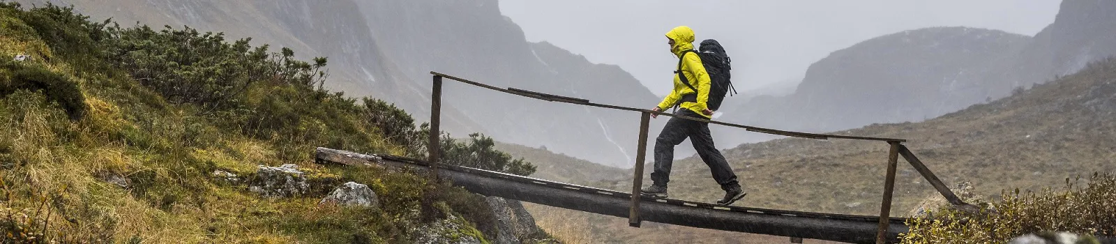Outdoor-Imagebild. Person mit gelber Jacke und Rucksck geht über einen kleinen Steg in den Bergen