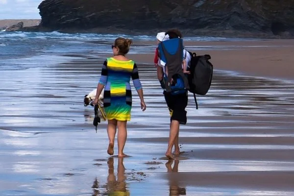 Outdoor Imagebild - zwei Personen laufen am Strand in flachem Wasser, eine Person trägt eine Kindertrage auf dem Rücken