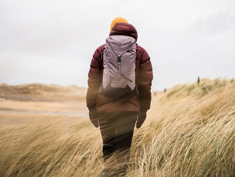 Outdoor Imagebild - Person mit Rucksack und Jacke ist von hinten zu sehen und läuft durch langes Gras.
