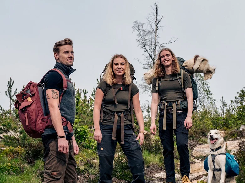 Outdoor Imagebild - drei Personen im freien mit Funktionsbekleidung und Rucksäcken. Ganz rechts am Bildrand ein Hund.