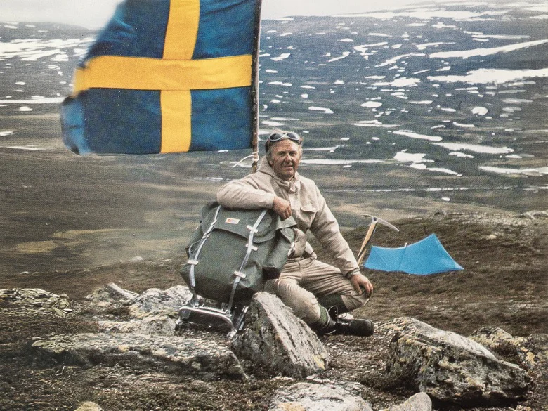 Outdoor Imagebild - Person sitzt auf einem Felsen in einer Tundra-artigen Landschaft, im Hintergrund ist eine schwedische Flagge zu sehen.